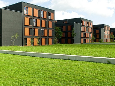 Wohnheim Stiftsbogen Auf dem Foto sieht man mehrere Neubauten und davor eine Rasenfläche. die Gebäude sind anthrazitfarben mit orangefarbenen Jalousien.
