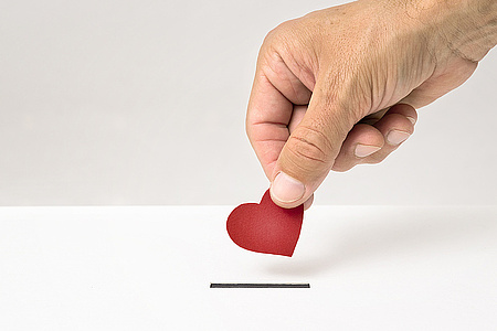 Eine Hand gibt ein kleines rotes Herz in den Schlitz einer Spendendose. 