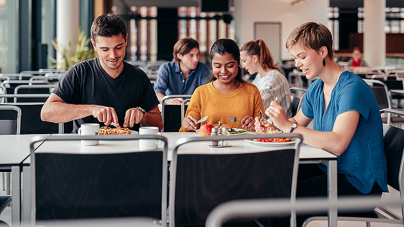 Drei Studierende sitzen an einem Tisch in der Mensa und Essen.