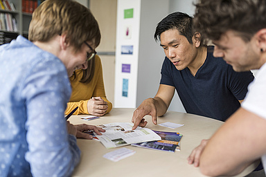 Drei Studierende sitzen gemeinsam mit einer Beraterin an einem Tisch und gehen Informationsmaterial durch.