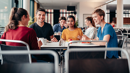 Drei junge Leute sitzend an einem Tisch in einer Mensa. Sie lächeln sich an. Vor ihnen stehen Becher und Teller. Eine der Personen hat eine Gabel in der Hand.