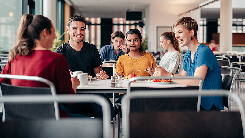 Drei junge Leute sitzend an einem Tisch in einer Mensa. Sie lächeln sich an. Vor ihnen stehen Becher und Teller. Eine der Personen hat eine Gabel in der Hand.