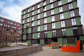 Holzgebäude mit abwechselnd dunkelgrün und anthrazitfarben angestichener Holzverkleidung, bodentiefe Fenster, davor Terrasse mit fest installierten Sitzmöbeln