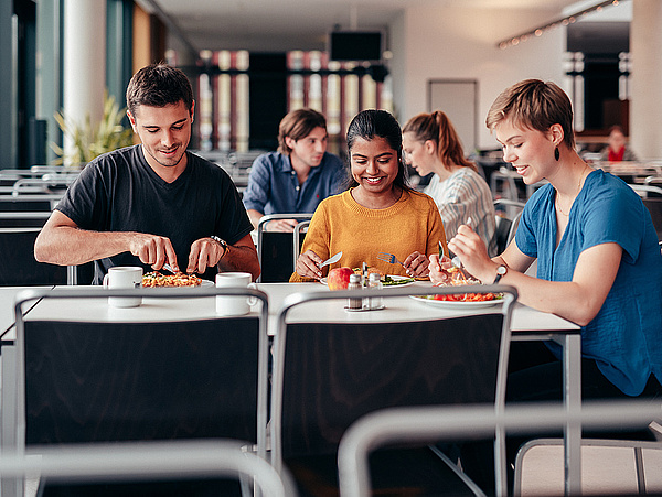 Drei Studiernede sitzen an einem Tisch in der Mensa und essen. 