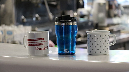 Zwei Tassen und ein Thermobecher stehen auf einer Theke. 