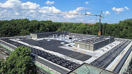 Auf dem Foto sieht man auf das Dach des Gebäudes der Mensa Leopoldstrasse. Es ist ein Flachdach. Dort wird etwas installiert - die PV-Anlage. Außerdem ist das Gebäude eingerüstet.