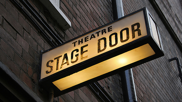 Symbolbild für Kultur: Backsteinhaus mit einem leuchtenden Bühnenschild, auf dem "Stage Door" steht.