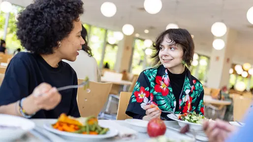 Zwei Studentinnen sitzen an einem Mensa Tisch und unterhalten sich beim Essen. Vor ihnen auf dem Tisch sind Teller und im linken Bildrand ist ersichtlich, dass ihnen auch jemand gegenüber sitzt. 