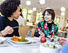 Zwei Studentinnen sitzen an einem Mensa Tisch und unterhalten sich beim Essen. Vor ihnen auf dem Tisch sind Teller und im linken Bildrand ist ersichtlich, dass ihnen auch jemand gegenüber sitzt. 