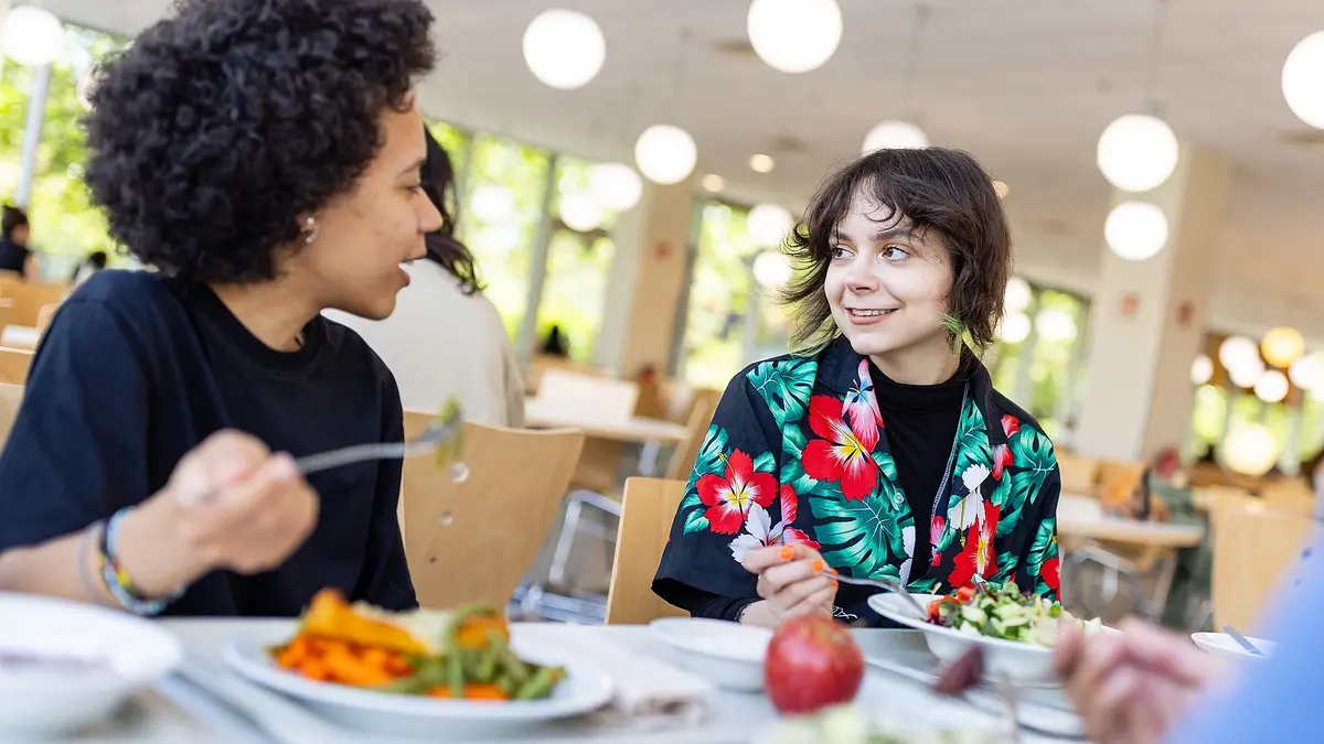 Zwei Studentinnen sitzen an einem Mensa Tisch und unterhalten sich beim Essen. Vor ihnen auf dem Tisch sind Teller und im linken Bildrand ist ersichtlich, dass ihnen auch jemand gegenüber sitzt. 