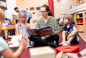 Zwei Kinder hocken neben einer Frau, die aus einem großen Buch vorliest.