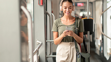 Frau mit Kopfhörern steht in einer Tram und blickt auf ihr Smartphone. 
