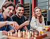 Vier Studierende sind auf dem Bild zu erkennen. Sie sitzen an einem Tisch und scheinen gute Stimmung zu haben. Zwei spielen Schach, auf dem Tisch stehen Getränke und Snacks. 