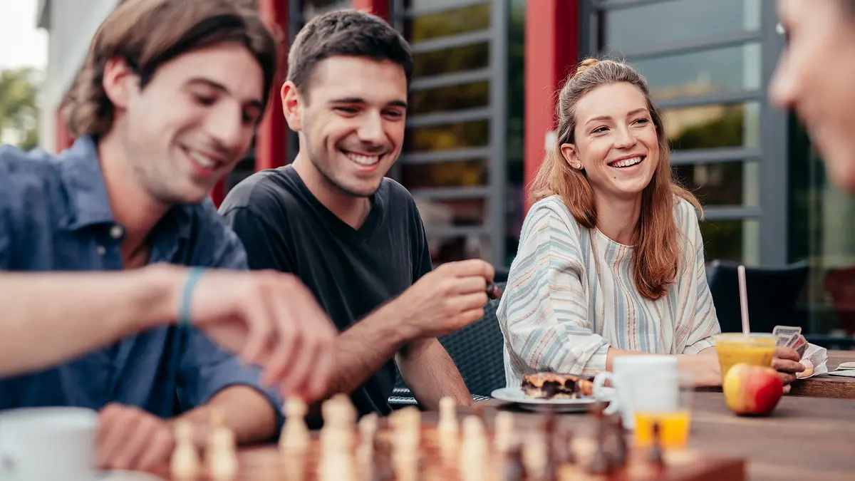 Vier Studierende sind auf dem Bild zu erkennen. Sie sitzen an einem Tisch und scheinen gute Stimmung zu haben. Zwei spielen Schach, auf dem Tisch stehen Getränke und Snacks. 