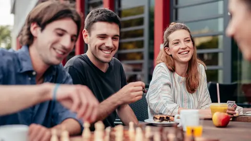 Vier Studierende sind auf dem Bild zu erkennen. Sie sitzen an einem Tisch und scheinen gute Stimmung zu haben. Zwei spielen Schach, auf dem Tisch stehen Getränke und Snacks. 