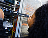 Nahaufnahme einer Person mit dunklem, lockigem Haar von der Seite, die eine flache Server-Einheit in ein Server-Rack hineinschiebt oder herauszieht. Im Hintergrund sind weitere Server-Komponenten, LED-Leuchten und verkabelte Netzwerkstrukturen in einem Rechenzentrum zu sehen.