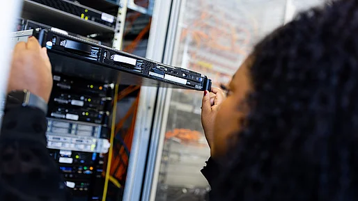 Nahaufnahme einer Person mit dunklem, lockigem Haar von der Seite, die eine flache Server-Einheit in ein Server-Rack hineinschiebt oder herauszieht. Im Hintergrund sind weitere Server-Komponenten, LED-Leuchten und verkabelte Netzwerkstrukturen in einem Rechenzentrum zu sehen.
