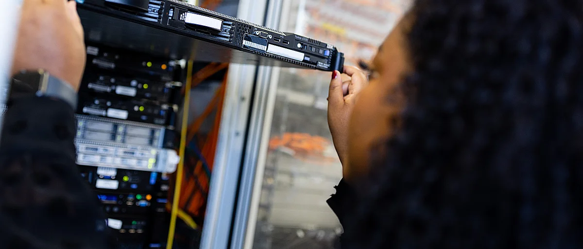 Nahaufnahme einer Person mit dunklem, lockigem Haar von der Seite, die eine flache Server-Einheit in ein Server-Rack hineinschiebt oder herauszieht. Im Hintergrund sind weitere Server-Komponenten, LED-Leuchten und verkabelte Netzwerkstrukturen in einem Rechenzentrum zu sehen.